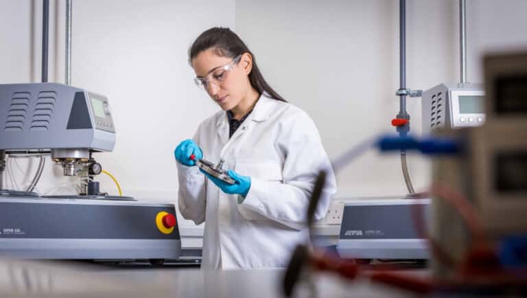 A person in a lab coat and safety goggles inspects a device in a laboratory setting with scientific equipment.