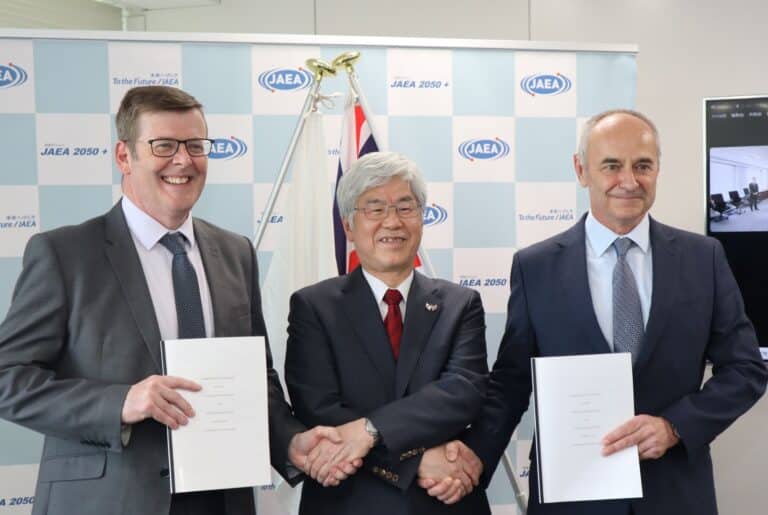 Three men in suits are smiling and shaking hands while holding documents, with a JAEA backdrop and a flag in the background.