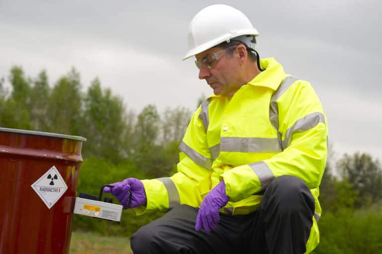 Person in safety gear uses a device to measure radiation near a marked barrel outdoors.